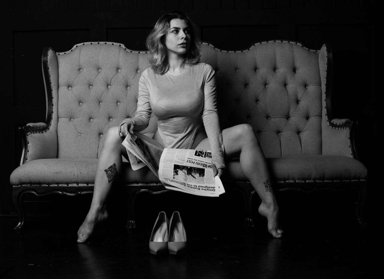 Stylish black and white portrait of a woman sitting on a sofa holding a newspaper.
