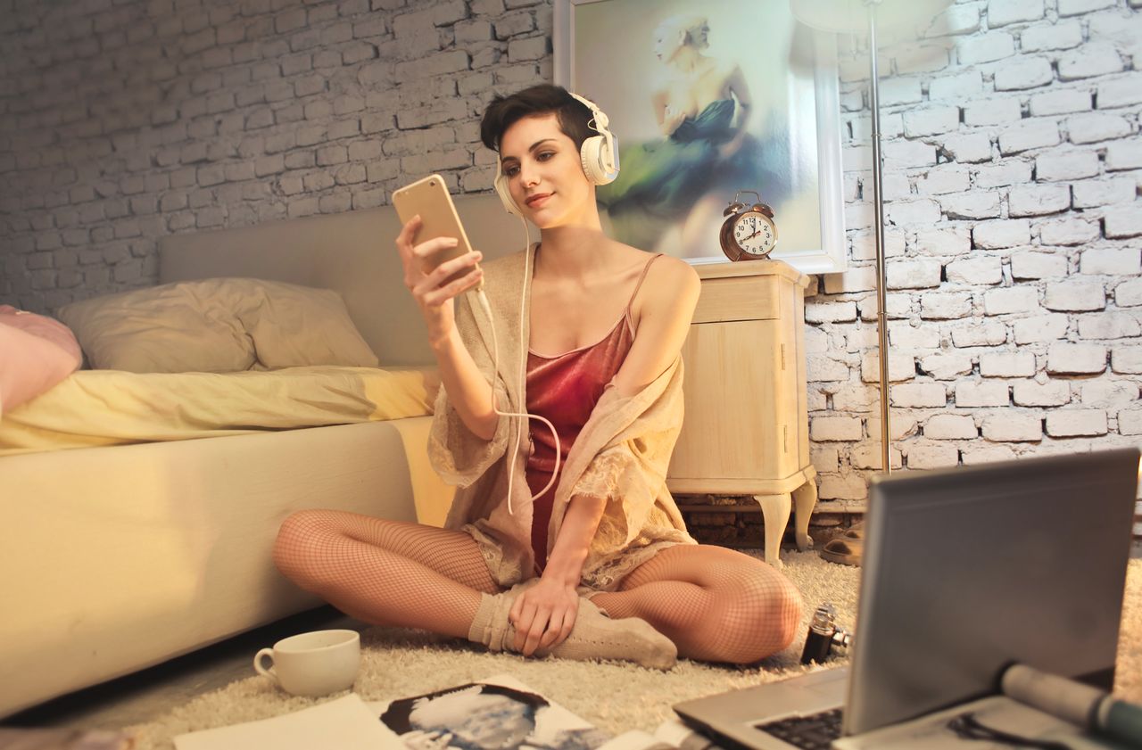 Young woman sitting on the floor with laptop and smartphone, enjoying music in a cozy bedroom.